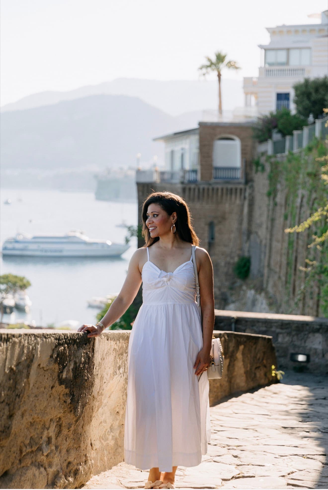Smiling woman in a white sundress overlooks a coastal harbor from a stone walkway.